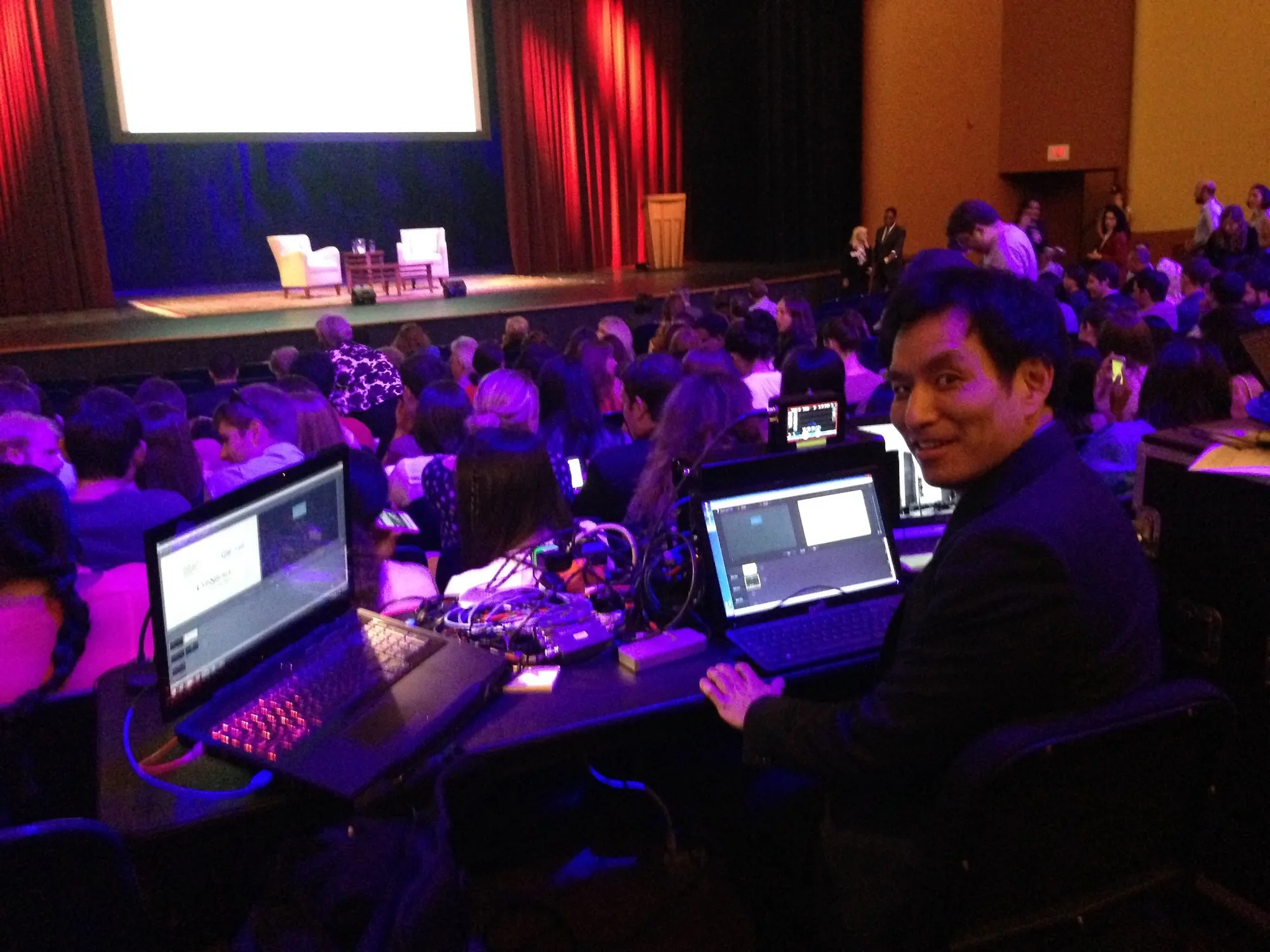 An Astrocasters operator at a laptop station in the audience of a theater event, with the stage and screen behind.