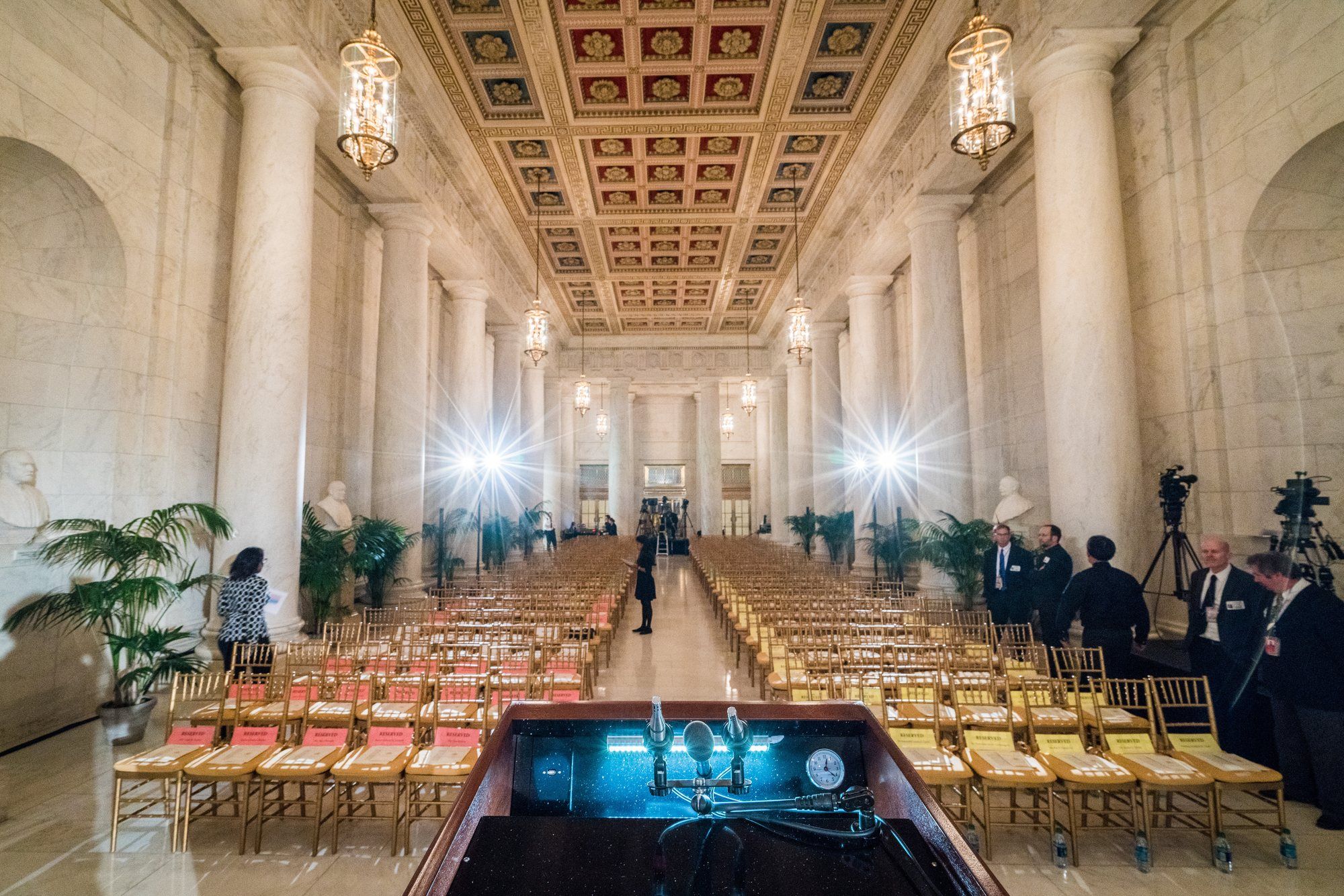 A long hall with marble columns set with rows of chairs, a lectern, lighting stands and cameras.