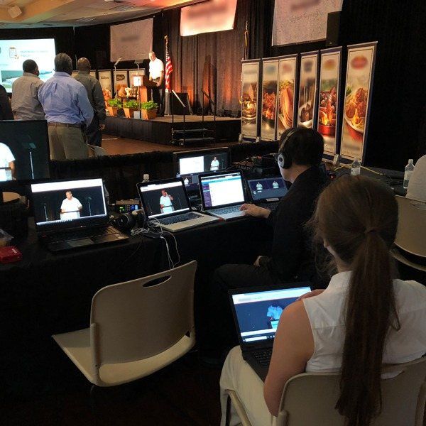 Operators with laptops and headsets at the back of a conference room, streaming the speaker at the lectern.
