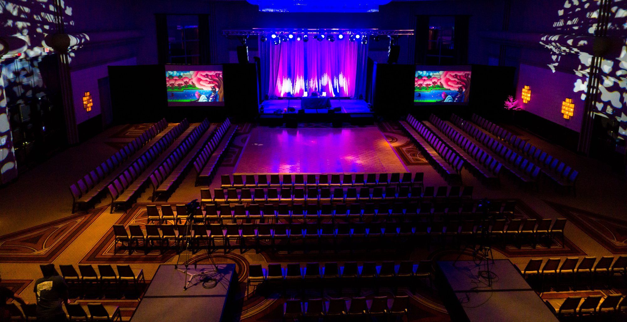A theater-style general session with a lit stage and a projection screen on each side.