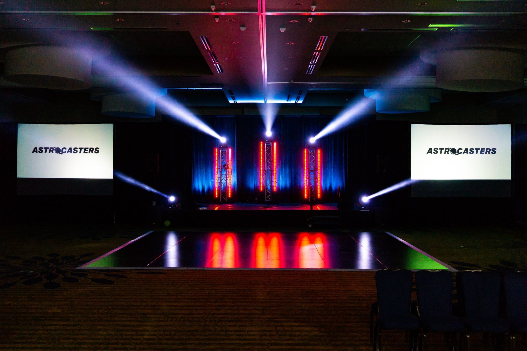 An event stage lit by moving-head lights in red and blue, with Astrocasters on the screens either side.